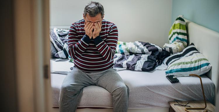 A man sitting on an unmade bed holding his hands over his face. 