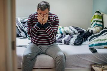 A man sitting on an unmade bed holding his hands over his face. 